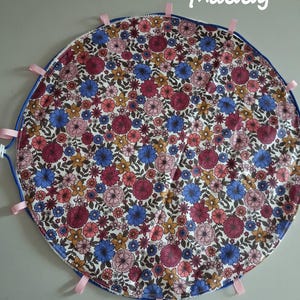 Op de afbeelding: Een ronde stoffen mat met een dicht bloemenpatroon in tinten blauw, roze en geel. De rand is afgewerkt met een blauwe bies en meerdere kleine, lichtroze stoffen lussen die naar buiten steken. De naam Maddy is in het wit gedrukt.