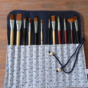 Può includere: Un rotolo di pennelli con stampa floreale blu con una sezione superiore a pois blu navy contiene vari pennelli. I pennelli hanno manici in legno e ghiera in metallo. Un cordoncino blu navy con perline di legno assicura il rotolo.