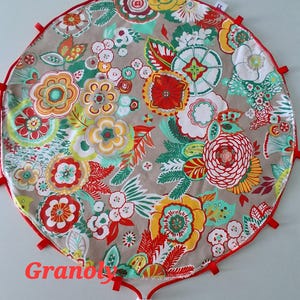 Op de afbeelding: Een ronde speelmat met bloemenpatroon, rode rand en strikjes. De mat heeft een beige achtergrond met kleurrijke bloemen in rood, geel, oranje en groen. Het woord "Granoly" is op de mat gedrukt.