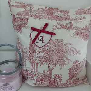 May include: A white pillow with a red toile pattern featuring a red ribbon tied in a bow around a heart-shaped embroidery with the letter "A" in the center.