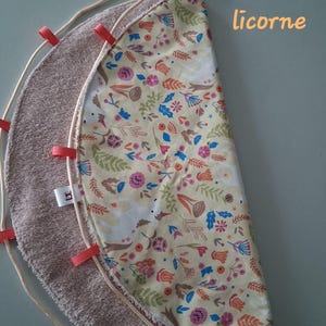 Op de afbeelding: Een halfronde speelmat met een lichtgele stoffen bovenkant met een kleurrijk bloemen- en eenhoornpatroon, een beige badstof onderkant en een rand afgezet met kleine oranje lintlussen en een dun koord. Het woord licorne is zichtbaar.