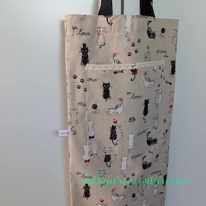 Può includere: Una borsa tote beige con manici neri e una stampa di gatti neri e bianchi. La borsa ha una piccola tasca con un bordo in pizzo e le parole "in love", "meow" e "purrr" sono stampate sulla borsa.