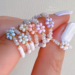 May include: A collection of beaded rings in various pastel colors, including blue, pink, and purple. Each ring features a flower design with gold accents. The rings are displayed on a person's finger, showcasing the delicate craftsmanship.