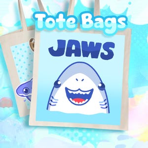 May include: Two beige canvas tote bags featuring a light blue square graphic. The front bag displays a cartoon shark with the text JAWS in dark blue letters above it. The shark has a wide, smiling mouth with visible teeth and dark eyes.