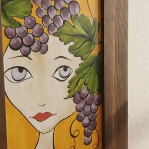 May include: A framed painting of a woman's face with a yellow background. The woman's hair is adorned with purple grapes and green leaves. The artwork has a whimsical style and is framed in a dark wood.