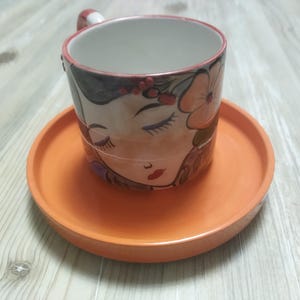 Könnte beinhalten: Ein Keramik-Tassen- und Untertassen-Set. Die Tasse zeigt ein gemaltes Gesicht mit geschlossenen Augen, floralen Akzenten und einer roten Lippe. Die Untertasse und der Henkel der Tasse sind orange. Die Tasse steht auf einer orangefarbenen Untertasse.