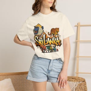 Op de afbeelding: Crèmekleurig T-shirt met een kleurrijk grafisch ontwerp met het woord "Sri Lanka". Het ontwerp bevat een aap, een luipaard en olifanten. Het shirt wordt gecombineerd met een spijkerbroekshort.