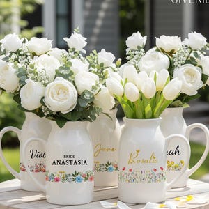 Op de afbeelding: Witte keramische kruiken met bloemstukken, waaronder pioenrozen en tulpen. De kruiken zijn gepersonaliseerd met namen en bloemmotieven. Het woord "GIVENIE" staat rechtsboven. De kruiken staan op een houten tafel.