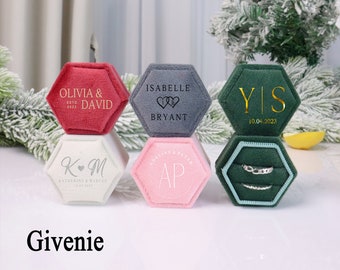 May include: Six hexagonal ring boxes with velvet interiors. The boxes are in various colors, including red, gray, green, white, and pink. Each box has a different inscription on the top, including "Olivia & David", "Isabelle Bryant", "YIS 10.04.2023", "KM Katherine & Marcus", and "AP".