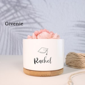 May include: White ceramic planter with a bamboo base. The planter has a black graduation cap and the name "Rachel" written in black cursive.