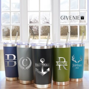 Peut inclure: Cinq gobelets en acier inoxydable personnalisés avec des pailles. Les gobelets sont de différentes couleurs : bleu, gris, noir, vert et turquoise. Chaque gobelet a un design différent, y compris une lettre, une ancre, une couronne et des bois de cerf. Les gobelets sont sur une surface en bois devant une fenêtre.