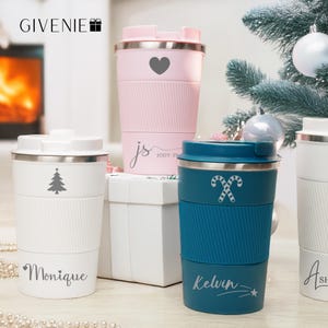 Puede incluir: Tres tazas de viaje personalizadas con diferentes diseños. La taza de la izquierda es blanca con un árbol de Navidad y el nombre "Monique". La taza del centro es azul con un diseño de bastón de caramelo y el nombre "Kelvin". La taza de la derecha es rosa con un corazón y el nombre "Jody".