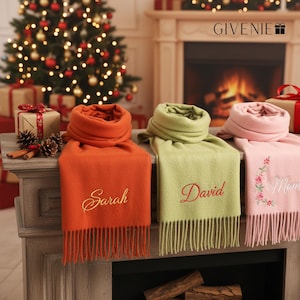 Könnte beinhalten: Drei personalisierte Schals in Orange, Grün und Rosa, mit gestickten Namen und floralen Details. Der orangefarbene Schal trägt den Namen "Sarah", der grüne Schal "David" und der rosa Schal "Mom". Jeder Schal hat einen Fransenrand. Weihnachtsbaum und Kamin im Hintergrund.