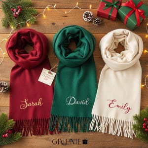 May include: Three personalized scarves in red, green, and cream, each with a name embroidered in a contrasting color. The scarves have fringed edges and are displayed on a wooden surface with Christmas decorations and gifts.