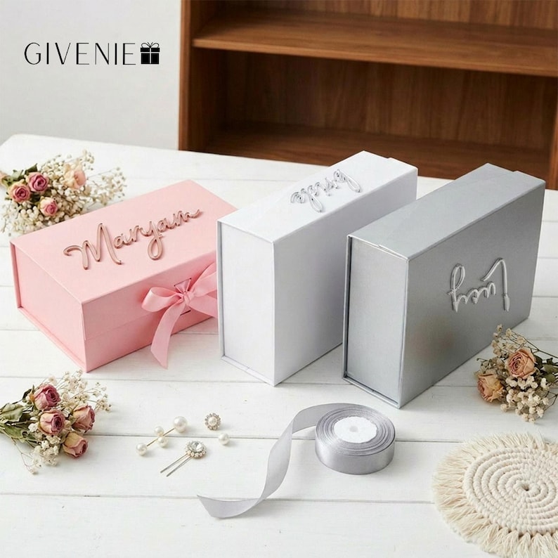 May include: Three rectangular gift boxes in pink, white, and silver, each with a name on top. The pink box has a matching ribbon. The boxes are on a white surface with flowers, jewelry, and a roll of ribbon.