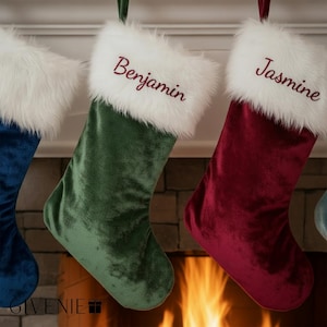 May include: Three velvet Christmas stockings in green, red, and blue hang from a fireplace mantel. Each stocking has a white faux fur cuff. The names "Benjamin" and "Jasmine" are embroidered on two of the stockings.