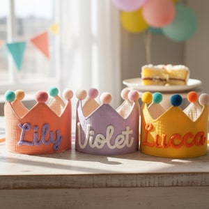 May include: Three colorful fabric crowns with pom-pom accents and personalized names: Lily, Violet, and Lucca. The crowns are in shades of orange, lavender, and yellow. The names are embroidered in a contrasting color. The crowns are sitting on a wooden surface.