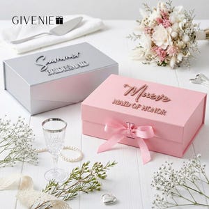 May include: Two gift boxes, one silver and one pink, are displayed. The silver box reads "Summer Bridesmaid" and the pink box reads "Maeve Maid of Honor". A pink ribbon adorns the pink box. The image includes a bouquet, a glass, and decorative elements.