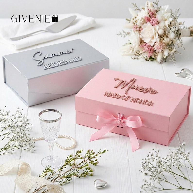 May include: Two gift boxes, one silver and one pink, are displayed. The silver box reads "Summer Bridesmaid" and the pink box reads "Maeve Maid of Honor". A pink ribbon adorns the pink box. The image includes a bouquet, a glass, and decorative elements.
