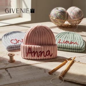 Könnte beinhalten: Drei gerippte Strickmützen in Hellblau, zartem Rosa und Salbeigrün, jeweils mit einem in dunkelrotem Schreibschriftgarn gestickten Namen. Die rosa Mütze ist mittig mit dem Namen Anna versehen, während die anderen Teilnamen oder Liam zeigen.