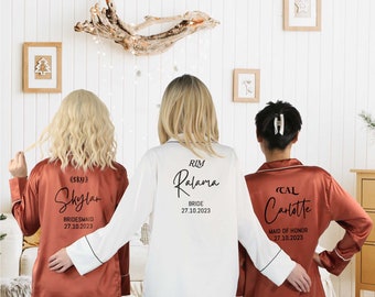 May include: Three matching satin pajamas with personalized text on the back. The pajamas are rust colored with black trim and white text. The text reads "Sky Skylar Bridesmaid 27.10.2023", "RIM Ralama Bride 27.10.2023", and "CAL Carlotte Maid of Honor 27.10.2023".