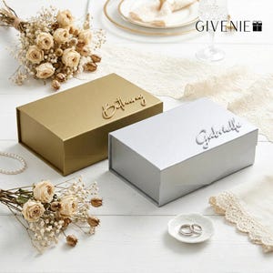 May include: Two rectangular gift boxes, one gold and one silver, sit on a white surface. The gold box has the name "Bethany" in cursive, while the silver box has "Gabrielle." Dried flowers and a small dish with rings are also present.