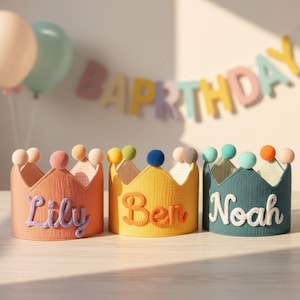 May include: Three colorful fabric crowns with the names "Lily", "Ben", and "Noah" embroidered on them. Each crown has a different color scheme, with pom-poms on top. The background includes balloons and a "Happy Birthday" banner.