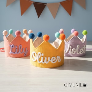 May include: Three colorful fabric crowns with the names "Lily", "Oliver", and "Violet" embroidered on them. Each crown is a different color: orange, yellow, and lavender. The crowns are decorated with colorful pom-poms and are likely for a child's birthday or party.