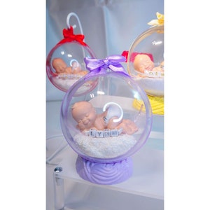 Op de afbeelding: Een transparante glazen bol met een paarse basis en een slapende babyfiguur erin. De baby is omringd door witte glitter en de naam "LYNN" is gespeld in witte letters.