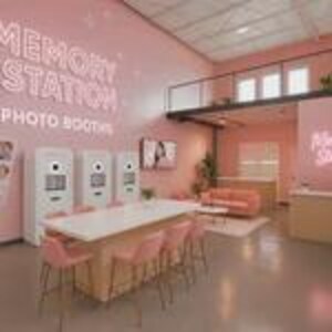 Op de afbeelding: Een roze fotostudio met drie witte fotohokjes, een lange witte tafel met roze stoelen en een roze bank. De woorden "MEMORY STATION PHOTO BOOTHS" staan in neonlicht op de muur. De studio heeft een mezzanine en een neutraal gekleurde balie.