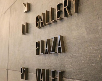 May include: A textured gray wall featuring three-dimensional dark bronze lettering. The text reads GALLERY PLAZA and VALET, accompanied by a small abstract logo symbol and the characters P1, arranged vertically in a clean, modern signage layout.