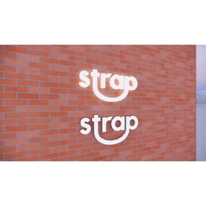 May include: A white sign with the word "strap" written twice on a brick wall. The sign is in a 3D style with a slight curve to the letters.