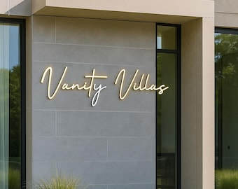 Può includere: Un'insegna a parete con il testo Vanity Villas in un carattere corsivo giallo luminoso. L'insegna è fissata su una facciata in pietra grigio chiaro con pannelli rettangolari, posizionata sopra un ingresso con finestre in vetro.