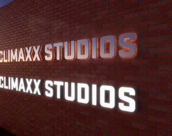 Può includere: Una parete in mattoni rossi con due insegne 3D illuminate. L'insegna superiore mostra il testo CLIMAXX STUDIOS con una finitura metallica e retroilluminazione arancione. L'insegna inferiore mostra il testo CLIMAXX STUDIOS in luce bianca.