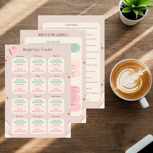 May include: A stack of three beige and pink weight loss tracker printables with the text "Weight Loss Tracker" and monthly sections. A cup of coffee with latte art and a small potted plant sit on a wooden surface.