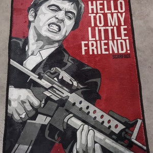 May include: A red and black doormat with a picture of a man holding a gun. The text "SAY HELLO TO MY LITTLE FRIEND! SCARFACE" is printed on the doormat.