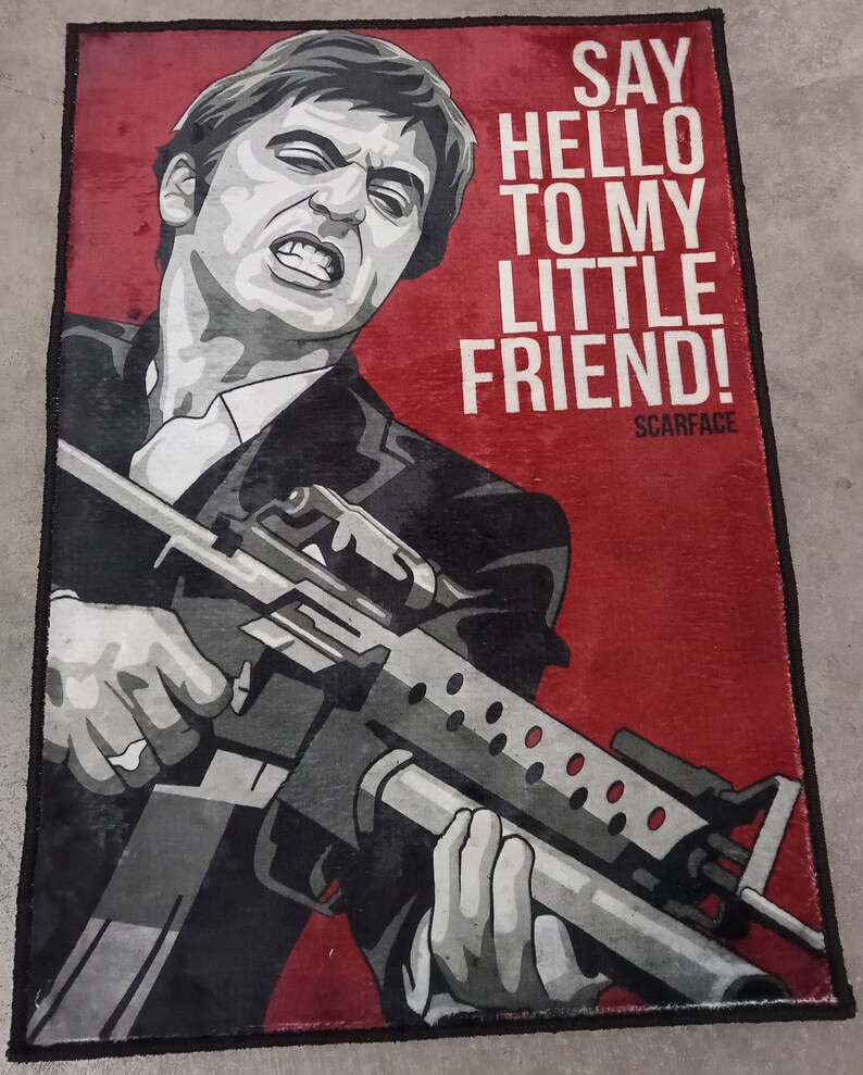 May include: A red and black doormat with a picture of a man holding a gun. The text "SAY HELLO TO MY LITTLE FRIEND! SCARFACE" is printed on the doormat.