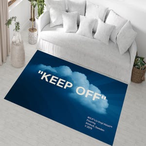 Può includere: Un tappeto rettangolare blu con un design a nuvola e le parole "KEEP OFF" in bianco. Il tappeto è posizionato davanti a un divano bianco. Il tappeto ha anche il testo "IKEA c/o Virgil Abloh™ Flooring Almhult, Sweden © 2019".