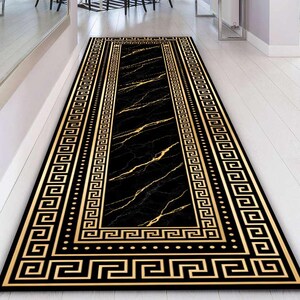 Può includere: Un tappeto runner rettangolare nero e oro con un bordo a chiave greca. Il centro presenta un motivo marmorizzato nero con venature dorate. Il tappeto è progettato per un corridoio o un ingresso.