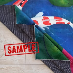 May include: A folded rug featuring a koi pond design with koi fish, lily pads, and a pink lotus flower. The rug has a blue, green, and white color scheme. The underside of the rug is black with white dots. The word "SAMPLE" is stamped on the rug.