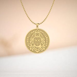 Puede incluir: Un collar con colgante dorado con un dije circular detallado. El dije muestra una cara tallada con patrones intrincados y un borde de cuentas. La cadena del collar es delicada y también dorada.