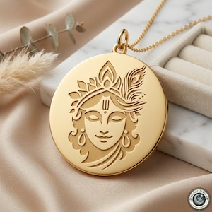 May include: A gold-colored pendant necklace featuring an engraved image of a serene face with ornate details, including a crown and feather. The round pendant hangs from a delicate gold chain, set against a neutral background.