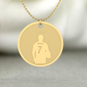 Puede incluir: Collar con colgante dorado. El colgante circular presenta la silueta de una persona que lleva una camiseta con el número 7 y el nombre "Ronaldo". El colgante cuelga de una delicada cadena dorada.