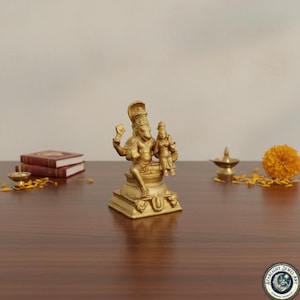 Può includere: Una statua dorata di una divinità con una figura seduta che tiene una figura più piccola. La statua è su una base quadrata con dettagli ornati. Altri oggetti includono libri, una piccola lampada e un fiore di calendula su una superficie di legno.