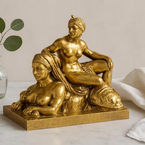 May include: A gold-colored sculpture depicting two figures, one reclining and the other seated above. The sculpture is on a rectangular base. A small glass vase with a green plant is to the left.