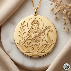 May include: A gold-colored pendant necklace featuring an engraved image of a seated figure playing a stringed instrument, with a swan and floral elements. The pendant is round and hangs from a delicate gold chain. The background is a marble surface with beige fabric.