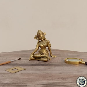 Puede incluir: Una estatua dorada de una figura femenina sentada con tocado y joyas ornamentadas. La estatua está sobre una superficie de madera, con un pincel, una pequeña plantilla de latón y una lupa cerca. La estatua es una representación de una deidad.