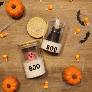 Könnte beinhalten: Halloween-Kerzen in Gläsern. Eine Kerze ist eine schwarze Katze, die andere ein rosa Geist. Beide Kerzen haben den Text "hey BOO" auf der Basis. Die Szene umfasst orangefarbene Kürbisse, Zuckermais und Fledermausdekorationen.