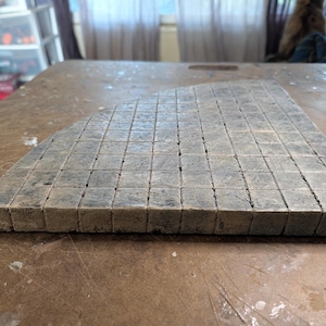 May include: A gray, triangular mosaic tile piece composed of numerous small, square tiles. The tiles have a weathered appearance, with variations in color and texture. The piece is set on a brown surface.