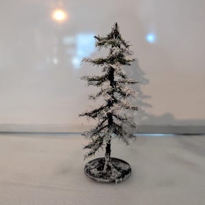 May include: A miniature model of a coniferous tree with dark green branches dusted with white flocking to resemble snow. The tree features a pointed top and is mounted on a small, circular, dark-coloured base that is also covered in white.